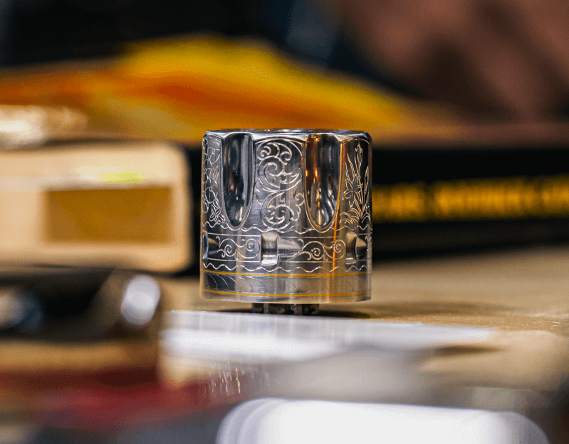 Close-up of a small, engraved metal cylinder resting on a tabletop, with blurred books and objects in the background.